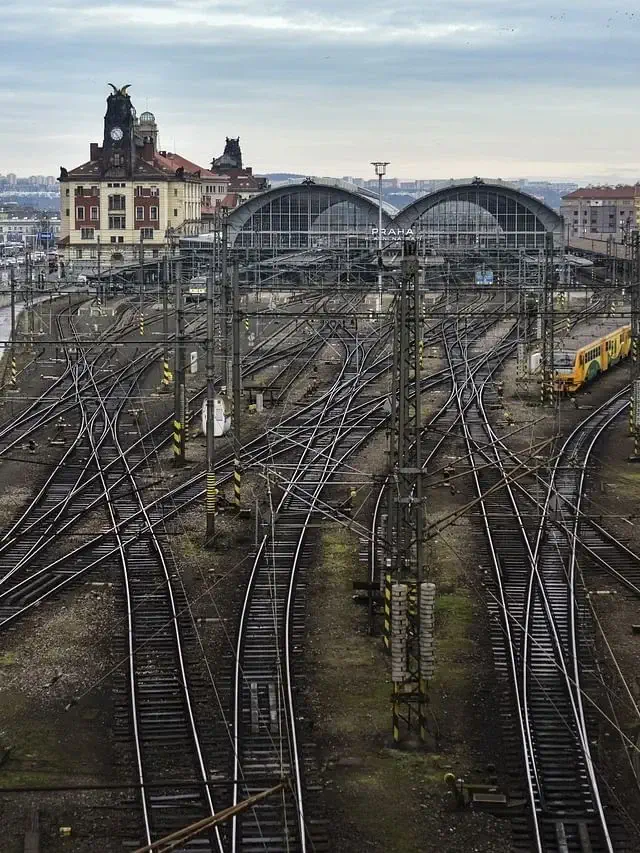 Mehrere Bahngleise mit Oberleitungen laufen zu einem historischen Bahnhof mit gewölbten Dächern und einem Uhrenturm zusammen - ein städtisches Wahrzeichen, das Reisende dazu einlädt, das All-Aboard-Paket zu erleben.